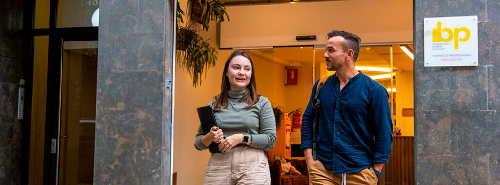 Two people are standing outside a building with a sign that reads "ibp International Business Programs Formació Professional Barcelona." The person on the left is holding a black folder and wearing a green long-sleeve shirt and beige pants. The person on the right is wearing a blue shirt and beige pants. The entrance to the building is visible behind them, with plants hanging on the wall and a fire extinguisher in the background.